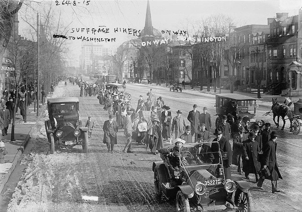 Photo shows the hike lead by "General" Rosalie Jones from New York to Washington, D.C. for the March 3, 1913 National American Woman Suffrage Association parade. Photo taken in Newark, New Jersey on Broad Street, just north of West Kinney Street, on February 12, 1913. Rosalie Jones is walking behind the first car. The church before City Hall is Grace Episcopal Churc
